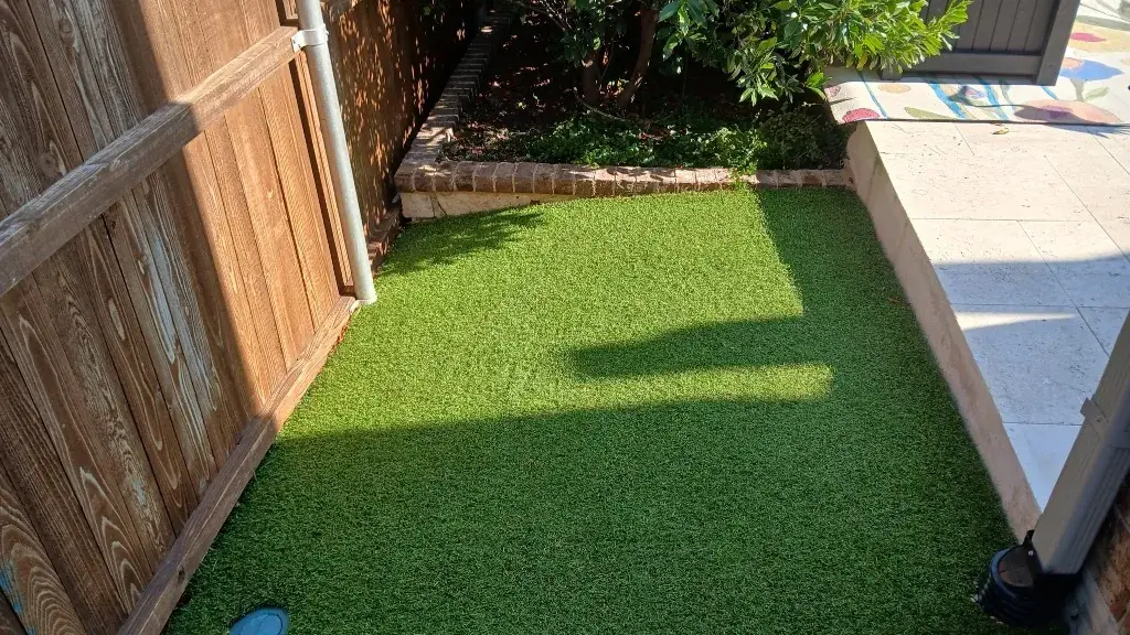 Side-yard turf installation bordered by a wood fence, brick planter wall, and a downspout tied into a drainage pipe